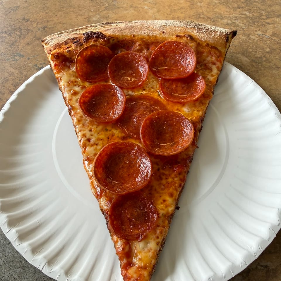 A large slice of pepperoni pizza with melted cheese and a crispy crust on a white paper plate.